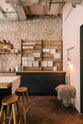 A quiet corner of a calm and simple Sebastian Cox kitchen features a long sink run with two sets of copper sinks and aged brass Mayan taps, a copper worktop, a shelf, and a prep table with a solid oak worktop. Adorned with vintage items, artisan ceramics, wooden chopping boards, tea towels, and wildflowers in large glass containers, this kitchen creates a chic New York industrial space that celebrates a love of natural materials.