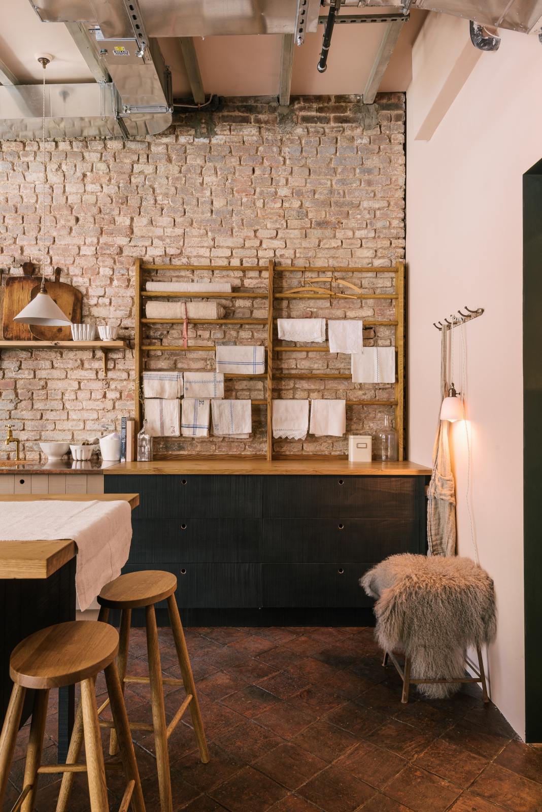 A quiet corner of a calm and simple Sebastian Cox kitchen features a long sink run with two sets of copper sinks and aged brass Mayan taps, a copper worktop, a shelf, and a prep table with a solid oak worktop. Adorned with vintage items, artisan ceramics, wooden chopping boards, tea towels, and wildflowers in large glass containers, this kitchen creates a chic New York industrial space that celebrates a love of natural materials.