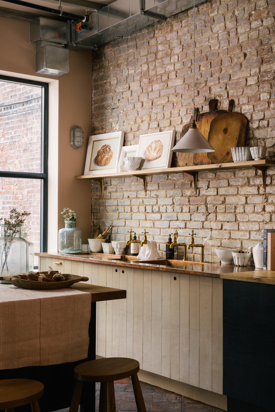 An angled view of a calm and simple Sebastian Cox kitchen features a long sink run with two sets of copper sinks and aged brass Mayan taps, a copper worktop, a shelf, and a prep table with a solid oak worktop. Adorned with vintage items, artisan ceramics, wooden chopping boards, framed illustrations, tea towels, and wildflowers in large glass containers, this kitchen creates a chic New York industrial space that celebrates a love of natural materials.