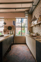 Nestled between the long sink run of a calm and simple Sebastian Cox kitchen, which features two sets of copper sinks and aged brass Mayan taps, a copper worktop, and a prep table with a solid oak worktop, this kitchen is adorned with vintage items, artisan ceramics, wooden chopping boards, framed illustrations, and a terracotta square floor. It creates a chic New York industrial space that celebrates a love of natural materials.