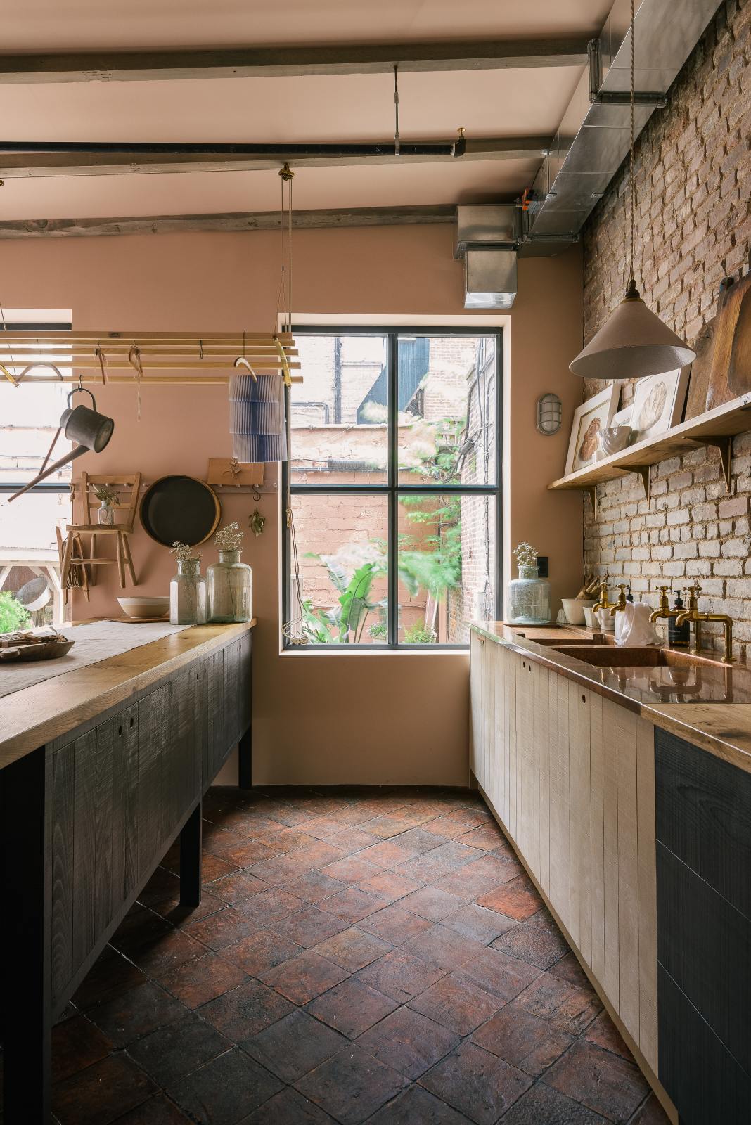 Nestled between the long sink run of a calm and simple Sebastian Cox kitchen, which features two sets of copper sinks and aged brass Mayan taps, a copper worktop, and a prep table with a solid oak worktop, this kitchen is adorned with vintage items, artisan ceramics, wooden chopping boards, framed illustrations, and a terracotta square floor. It creates a chic New York industrial space that celebrates a love of natural materials.