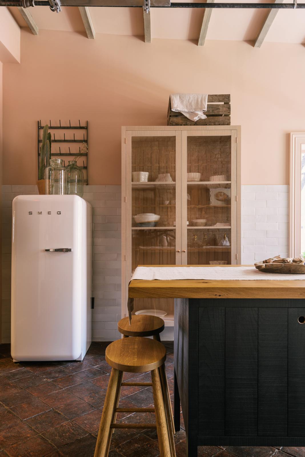 A quiet corner of a calm and simple Sebastian Cox kitchen features a long sink run with two sets of copper sinks and aged brass Mayan taps, a copper worktop, a shelf, and a prep table with a solid oak worktop. Adorned with vintage items, artisan ceramics, chopping boards, tea towels, and wildflowers in large glass containers, this kitchen creates a chic New York industrial space that celebrates a love of natural materials.
