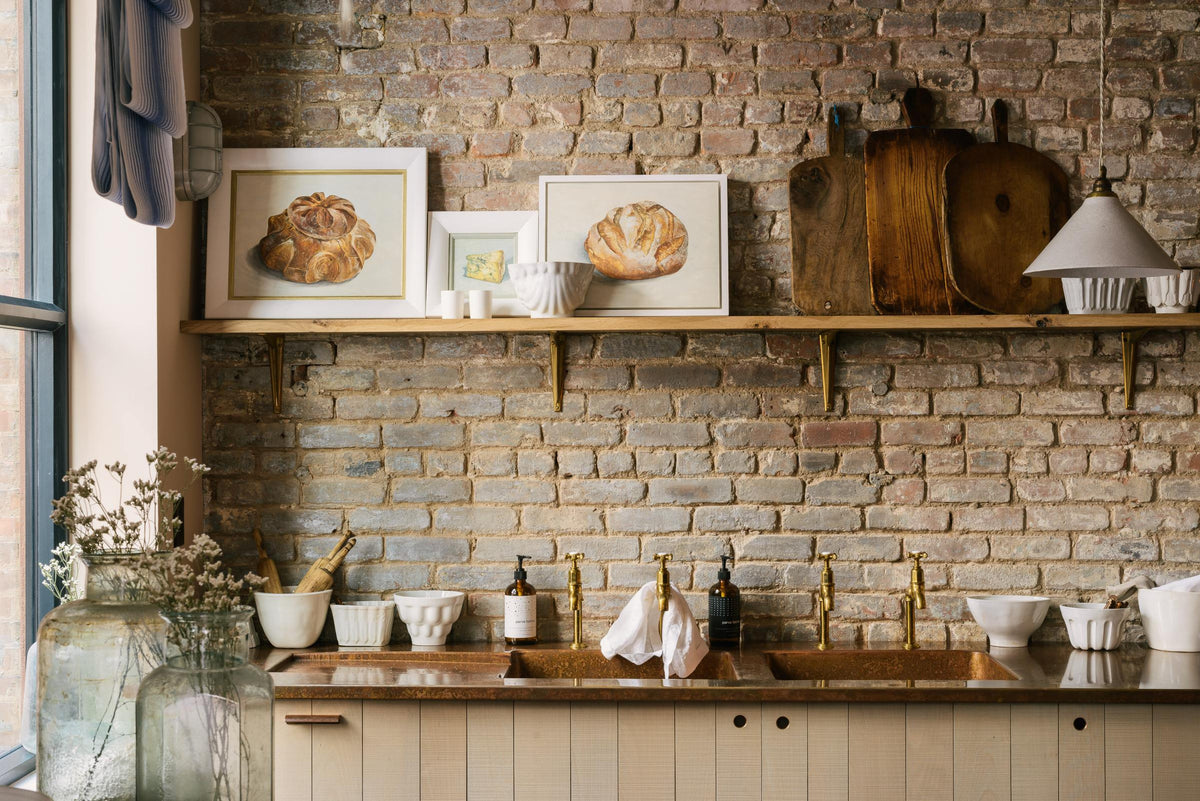 The other side of a calm and simple Sebastian Cox kitchen features a prep table with a solid oak worktop, a breakfast nook, a glazed freestanding cupboard, and a large white refrigerator. Adorned with vintage items, artisan ceramics, a cactus plant, framed illustrations, tea towels, and Helen stools, this kitchen creates a chic New York industrial space that celebrates a love of natural materials.