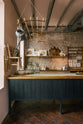 A calm and simple Sebastian Cox kitchen features a long sink run with two sets of copper sinks and aged brass Mayan taps, a copper worktop, a shelf, and a prep table with a solid oak worktop. Adorned with vintage items, artisan ceramics, wooden chopping boards, framed illustrations, tea towels, and wildflowers in large glass containers, this kitchen creates a chic New York industrial space that celebrates a love of natural materials.