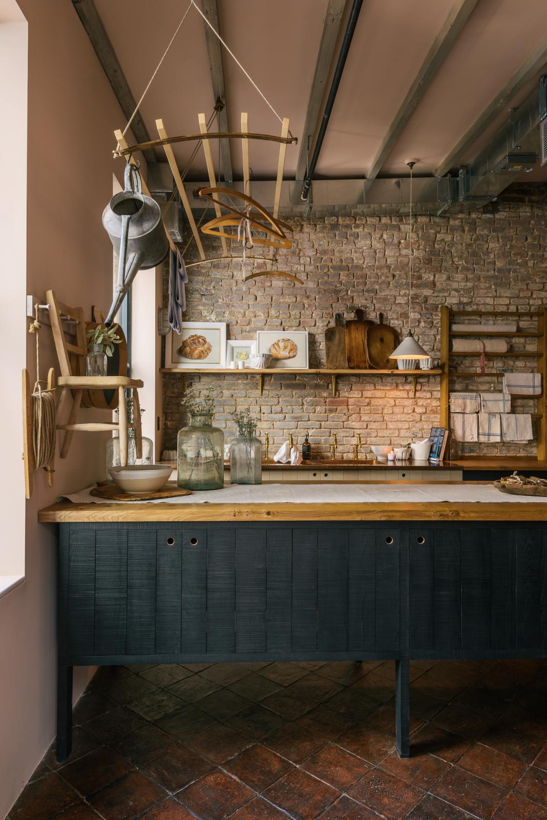 A calm and simple Sebastian Cox kitchen features a long sink run with two sets of copper sinks and aged brass Mayan taps, a copper worktop, a shelf, and a prep table with a solid oak worktop. Adorned with vintage items, artisan ceramics, wooden chopping boards, framed illustrations, tea towels, and wildflowers in large glass containers, this kitchen creates a chic New York industrial space that celebrates a love of natural materials.