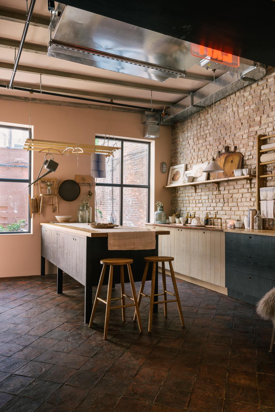 An oblique view of a calm and simple Sebastian Cox kitchen features a long sink run with two sets of copper sinks and aged brass Mayan taps, a copper worktop, a shelf, and a prep table with a solid oak worktop. Adorned with vintage items, artisan ceramics, framed illustrations, tea towels, a creamware pendant light, and a deVOL laundry maid, this kitchen creates a chic New York industrial space that celebrates a love of natural materials.