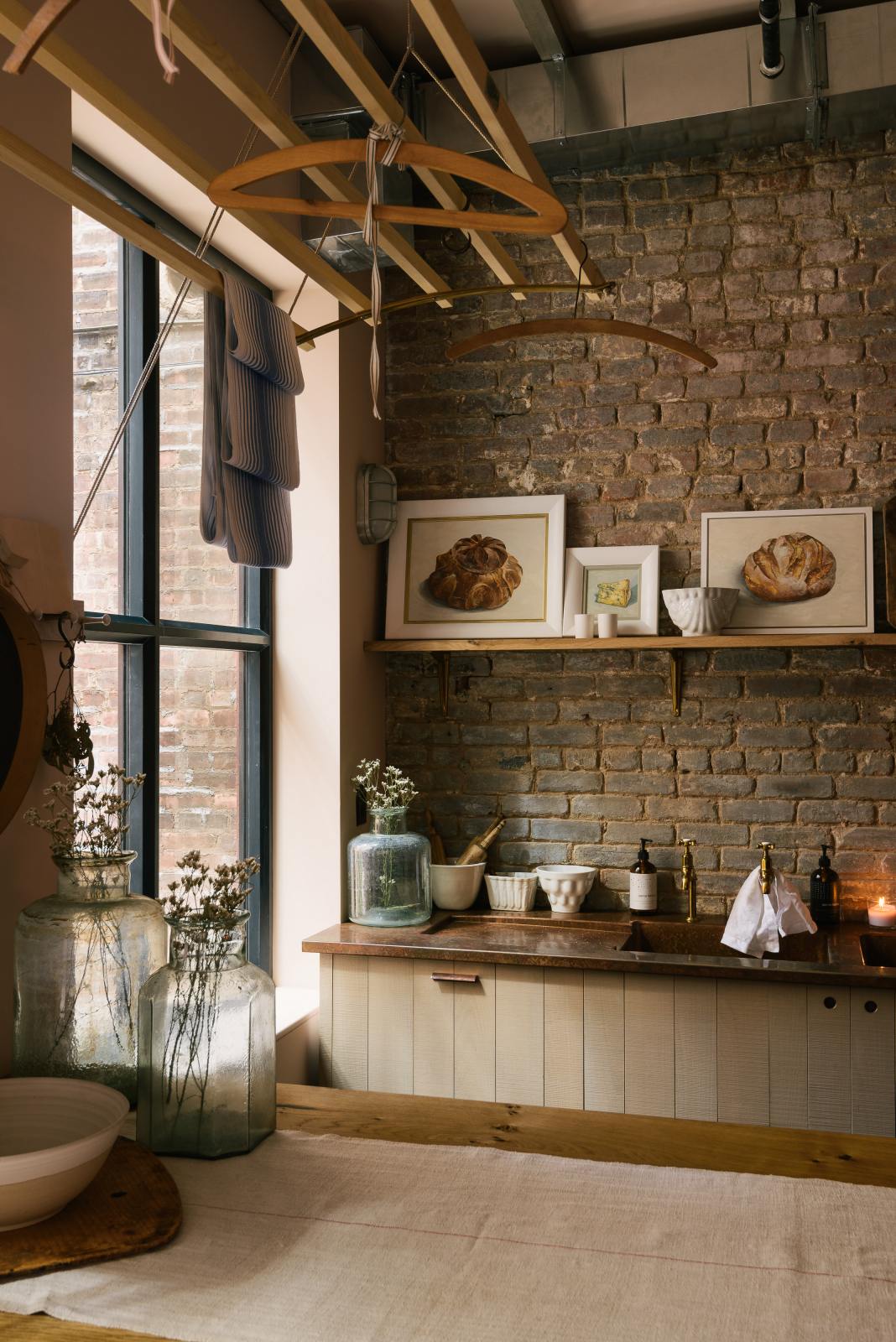 A calm and simple Sebastian Cox kitchen features a long sink run with two sets of copper sinks and aged brass Mayan taps, a copper worktop, a shelf, and a prep table with a solid oak worktop. Adorned with vintage items, artisan ceramics, wooden chopping boards, framed illustrations, tea towels, and wildflowers in large glass containers, this kitchen creates a chic New York industrial space that celebrates a love of natural materials.