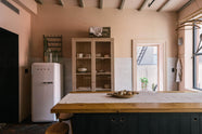 A calm and simple Sebastian Cox kitchen features a prep table with a solid oak worktop, and a glazed freestanding cupboard. Adorned with vintage items, artisan ceramics, wooden chopping boards, framed illustrations, tea towels, and wildflowers in large glass containers, this kitchen creates a chic New York industrial space that celebrates a love of natural materials.