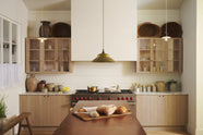 An all-natural and functional Sebastian Cox kitchen features a cooker and sink run with a marble worktop, glazed wall cupboards, and an iconic Haberdasher’s prep table with a copper worktop. Lovingly adorned with antique artisan ceramics, woven baskets, beautiful houseplants, and beloved figurines, it creates a wonderfully simple kitchen with a love of natural sustainability. 