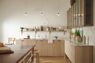 An all-natural and functional Sebastian Cox kitchen features an L-shaped sink run with a marble worktop, glazed wall cupboards, and an iconic Haberdasher’s prep table with a copper worktop. Lovingly adorned with antique artisan pottery, ceramic pendant lights, beautiful houseplants, and beloved figurines, it creates a wonderfully simple kitchen with a love of natural sustainability.