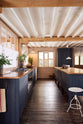 Nestled between the sink run of a warm and stylish Sebastian Cox kitchen, which features two single copper sinks, aged brass Mayan taps, a copper worktop, and a large prep table with a solid oak worktop, this kitchen is adorned with an aged brass café curtain rail, a milk glass task light, a creamware rise-and-fall light, artisan ceramics, and vintage wooden items. It creates a humble space that celebrates a love of simple and natural materials.