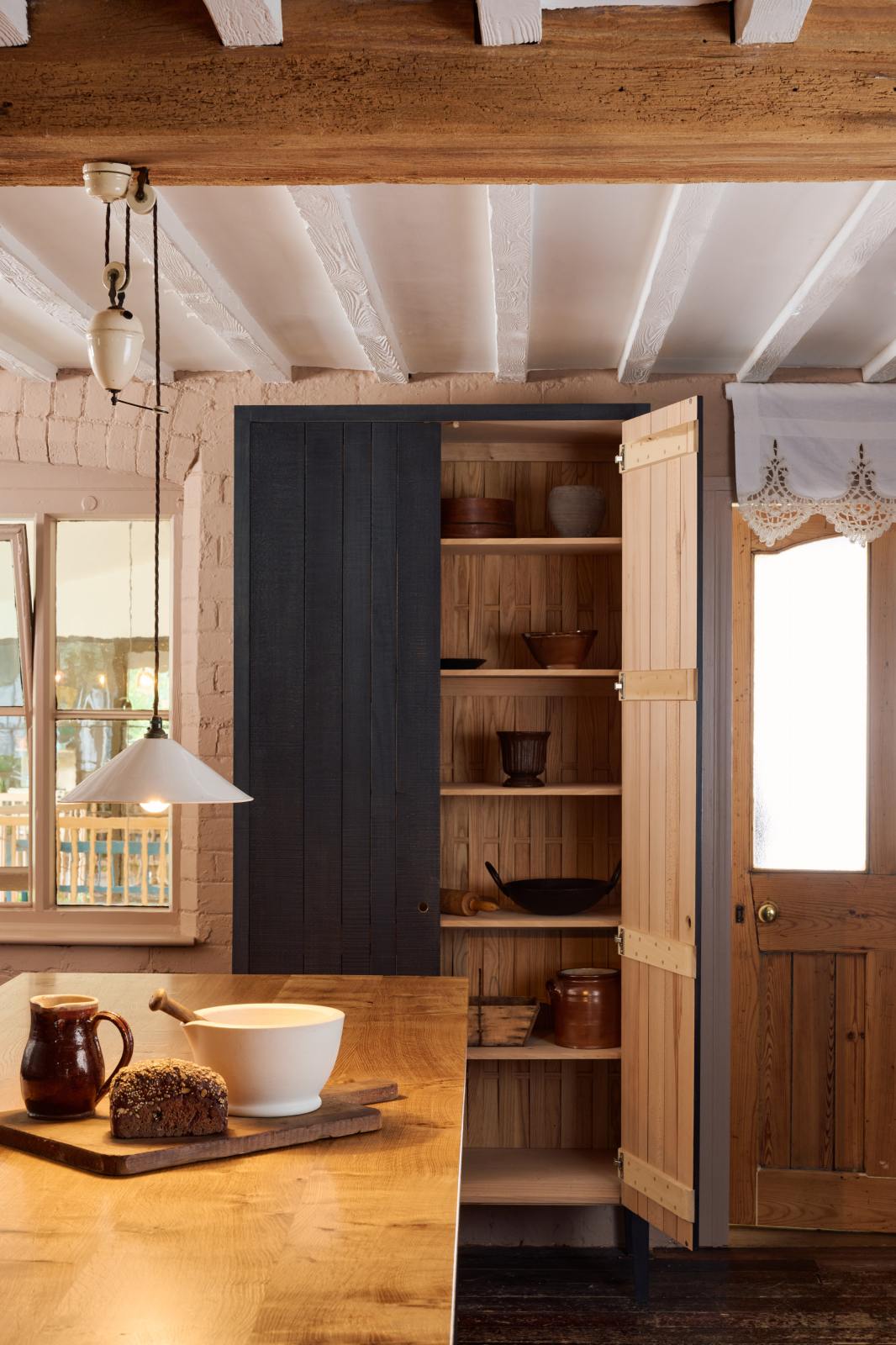 A warm and stylish Sebastian Cox kitchen features a prep table with a solid oak worktop and a freestanding cupboard with built-in shelves. Adorned with a creamware rise and fall, artisan ceramics, and vintage wooden items, this kitchen creates a humble space that celebrates a love of simple and natural materials.