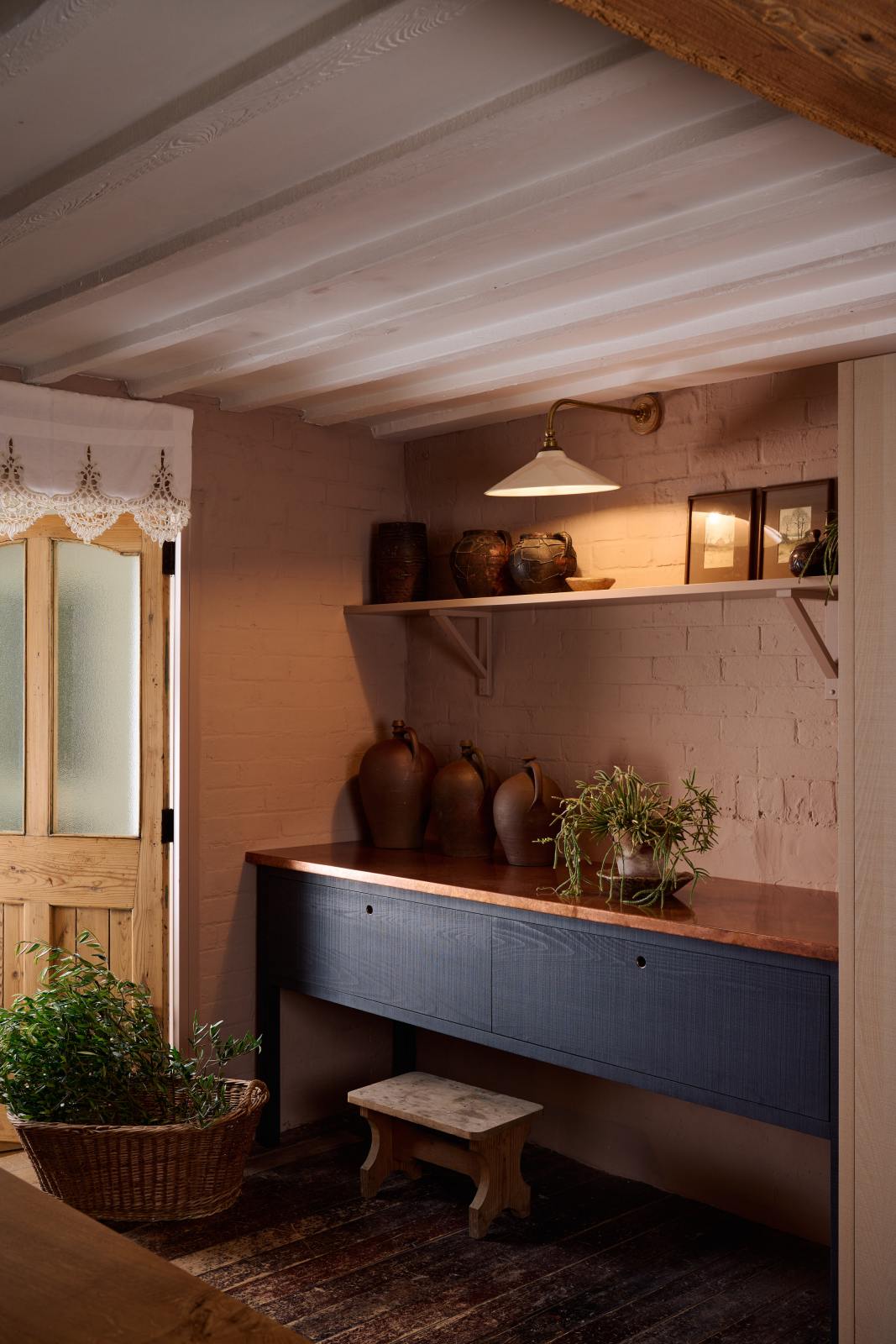 An oblique view of a warm and stylish Sebastian Cox kitchen features a small prep table with a copper worktop and a bank of free-standing cupboards. Adorned with a milk glass task light, artisan ceramics, and vintage wooden items, this kitchen creates a humble space that celebrates a love of simple and natural materials.
