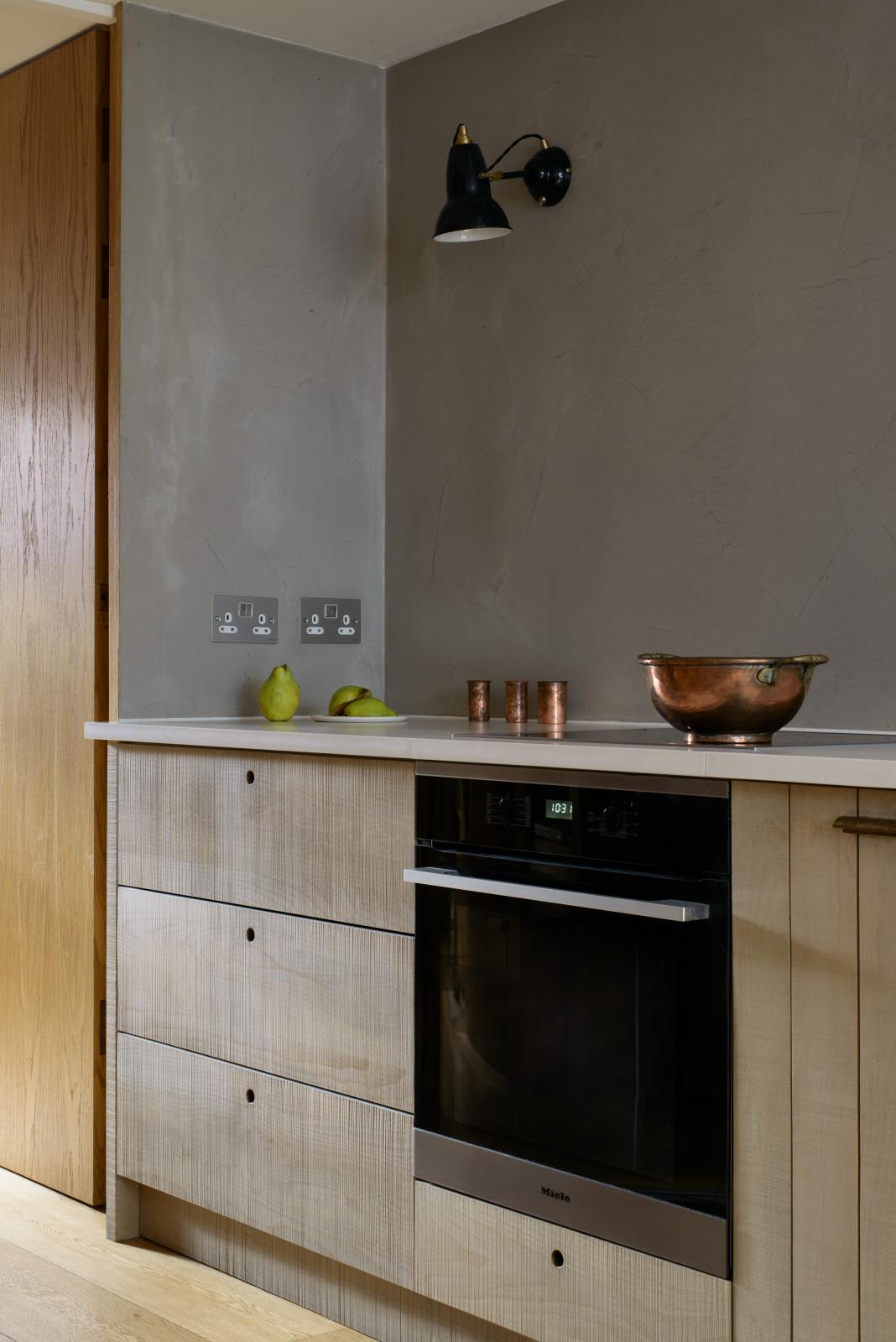 An oblique view of a sleek and simple Sebastian Cox kitchen includes a cooker and sink run topped with a Silestone worktop, and walls finished with natural limestone. The space is beautifully decorated with a plate of fresh green pears, copper shot glasses, a copper bowl, and a black industrial wall lamp, creating a minimal yet earth-conscious and sustainable kitchen environment.