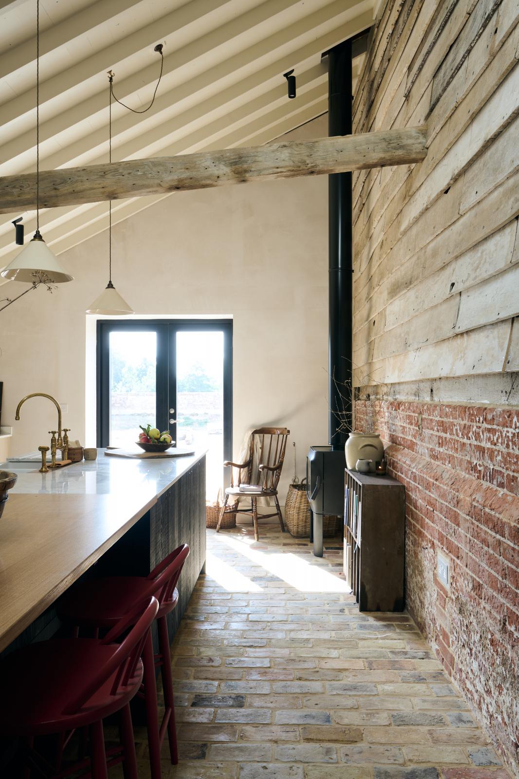 At the edge of a beautifully renovated farm, it features a Sebastian Cox kitchen and a cosy reading nook with a small burning wood cast iron fireplace, bookshelves, a vintage chair, and a basket of apples on a rustic original stone floor, along with exposed brickwork and wooden walls.