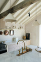 A close and oblique view of a beautifully renovated farm features a Sebastian Cox kitchen with a long cooker run, a large kitchen island with a double fluted Belfast sink, an aged brass Ionian tap, and a Carrara marble worktop. Adorned with creamware pendant lights, Heirloom wall lights, vintage items, and artisan ceramics, this kitchen creates a clean and contemporary style that harmonises with the natural materials used throughout the space.