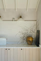 A quiet corner of a beautifully renovated farm features a Sebastian Cox kitchen with a long cooker run, a large kitchen island with a double fluted Belfast sink, an aged brass Ionian tap, and a Carrara marble and solid oak worktop. Adorned with Heirloom wall lights, vintage items, artisan ceramics, and an oil painting, this kitchen creates a clean and contemporary style that harmonises with the natural materials used throughout the space.