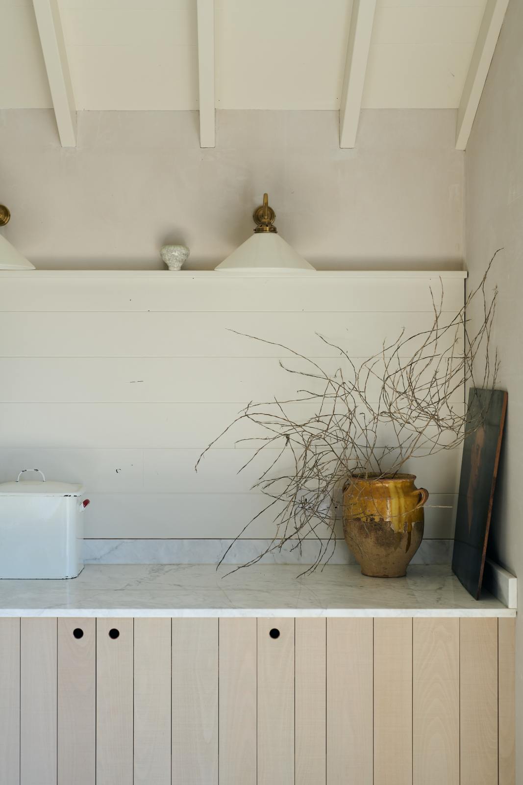 A quiet corner of a beautifully renovated farm features a Sebastian Cox kitchen with a long cooker run, a large kitchen island with a double fluted Belfast sink, an aged brass Ionian tap, and a Carrara marble and solid oak worktop. Adorned with Heirloom wall lights, vintage items, artisan ceramics, and an oil painting, this kitchen creates a clean and contemporary style that harmonises with the natural materials used throughout the space.
