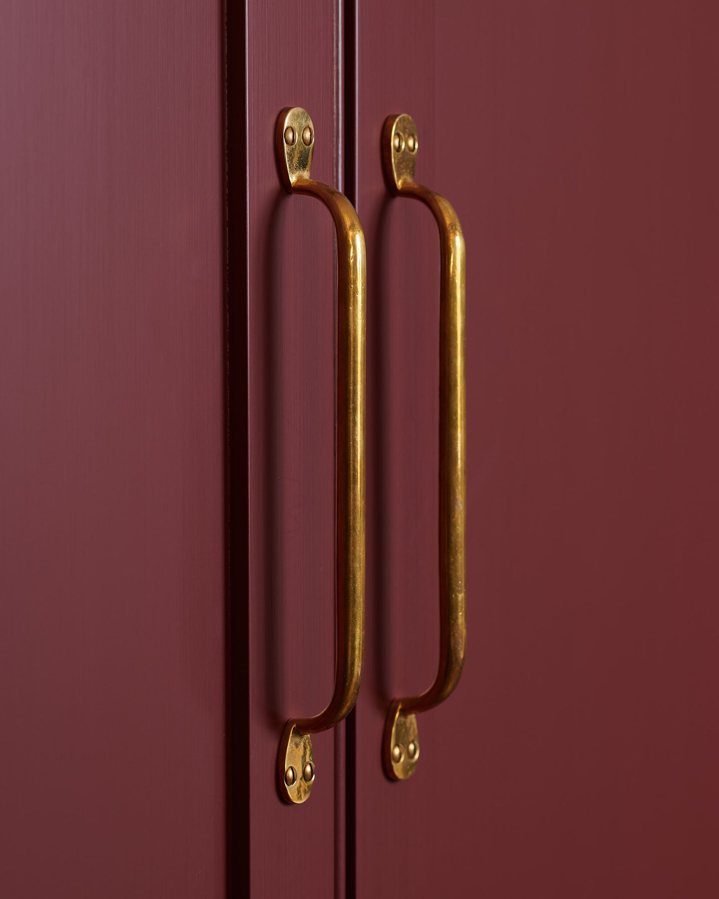 A close-up oblique view of a pair of classic pull handles in a warm burnished brass finish fitted to pantry doors painted in a wine-red colour.