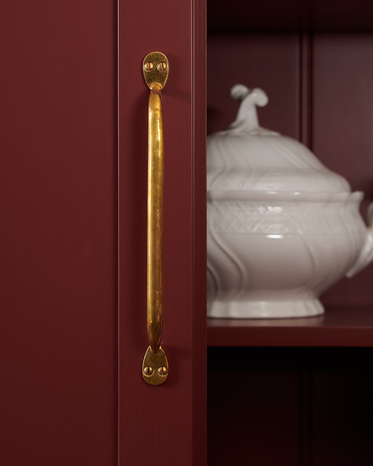 A close-up detailed shot of a large deVOL ‘Pull Handle’, fitted to the door of a maroon-coloured pantry cupboard. The golden ‘Heirloom Brass’ finish reflects the warm tones of the cupboard to create a subtle glow.