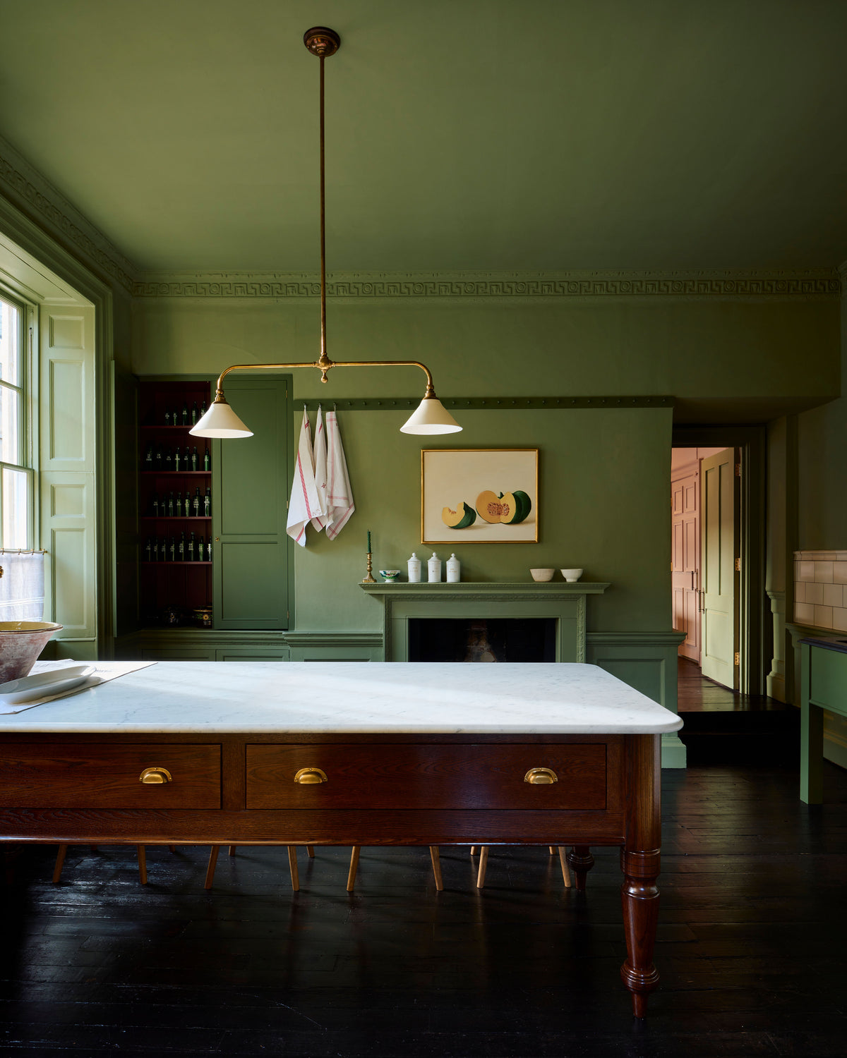 An atmospheric room with dark green walls and ceiling, black floorboards and a Victorian-inspired chandelier light with brass arms and small unglazed porcelain shades over a large oak prep table with a Carrara marble worktop.