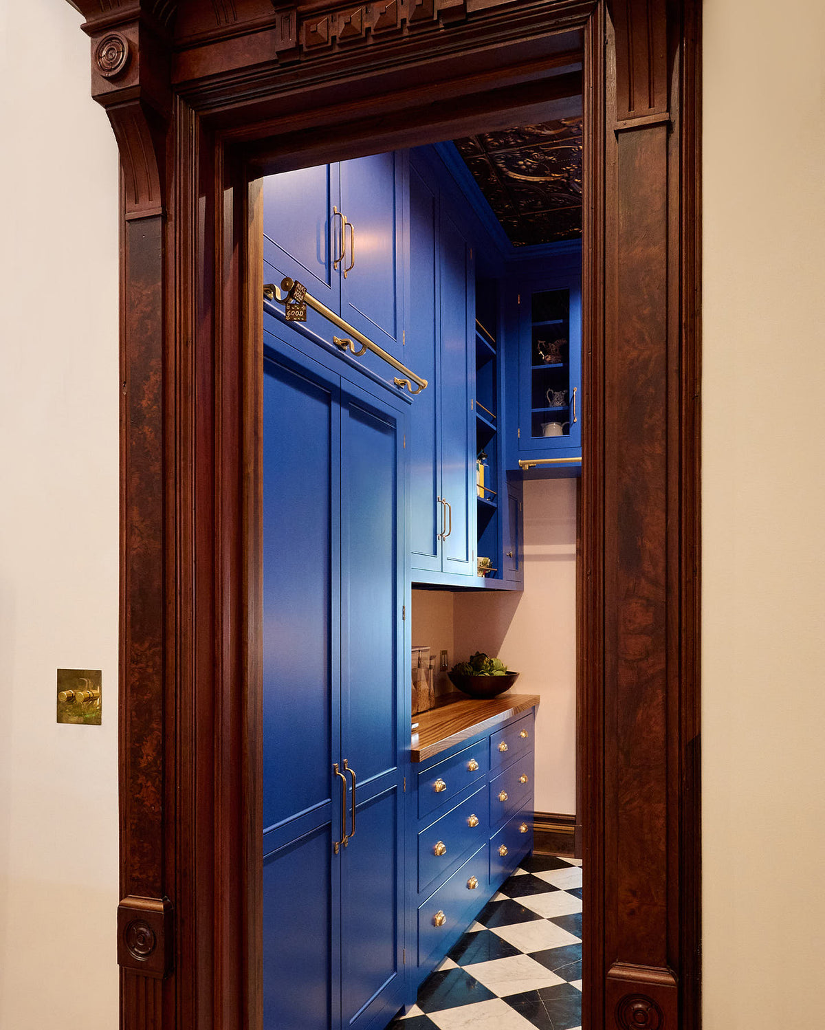 Beyond an ornate wooden doorway lies a walk-in galley pantry that boasts full-height cupboards, wall cupboards, and a drawer base cabinet topped with solid oak. This pantry is beautifully decorated with artisan ceramics, a bowl of artichokes, and dried ingredients stored in clear containers, all set against checkered marble tiles. The thoughtful design creates a social space that exudes a touch of luxury, making it perfect for both cooking and entertaining.