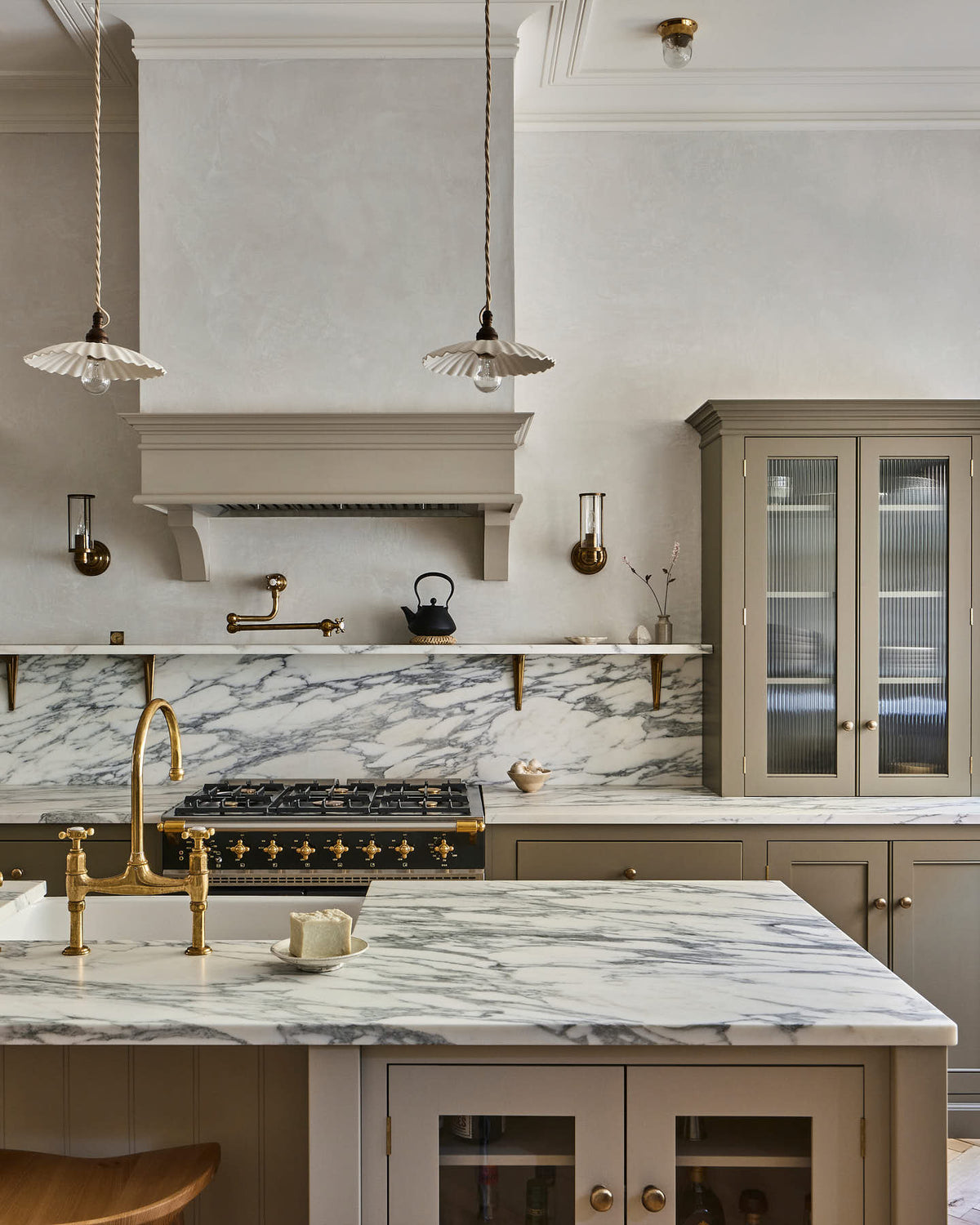 An elegant mushroom-brown, classic English kitchen features a cooker run with an Arabescato marble worktop, splash back, and shelf; a reeded countertop cupboard; a full-height pantry cupboard; and a large kitchen island with a fluted Belfast sink and an aged brass Ionian tap. Beautifully decorated with artisan ceramics, narrow pleated porcelain lights, deVOL Bum Stools, and a cast-iron teapot, it adds a sophisticated touch to the elegant kitchen.