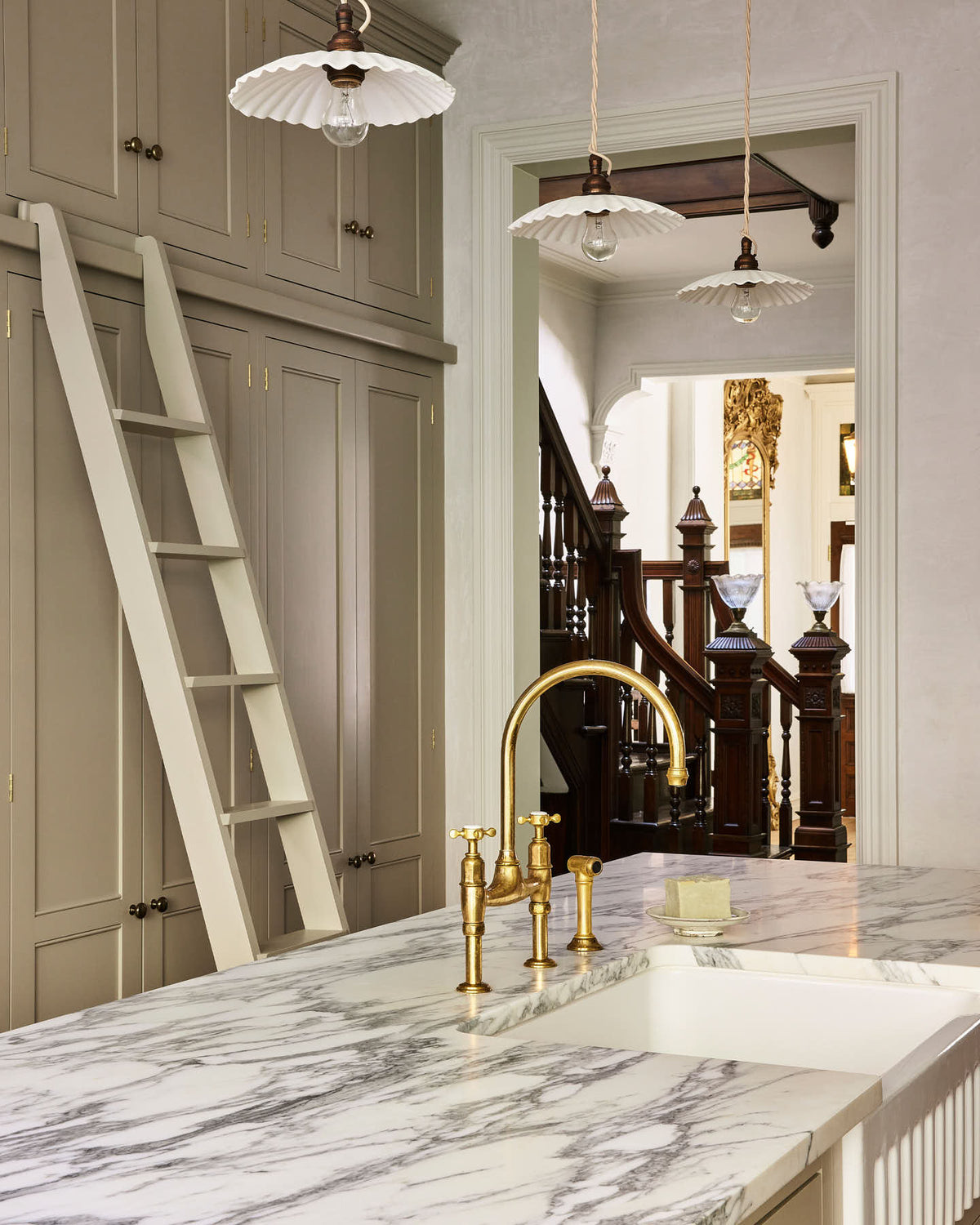 An angled look at an elegant mushroom-brown, classic English kitchen features a large kitchen island with a fluted Belfast sink, an aged brass Ionian tap, Arabescato marble worktop, and a full-height pantry cupboard with a ladder. Beautifully decorated with artisan ceramic, narrow pleated porcelain lights, and a bar of hand-made soap, it adds a sophisticated touch to the elegant kitchen.