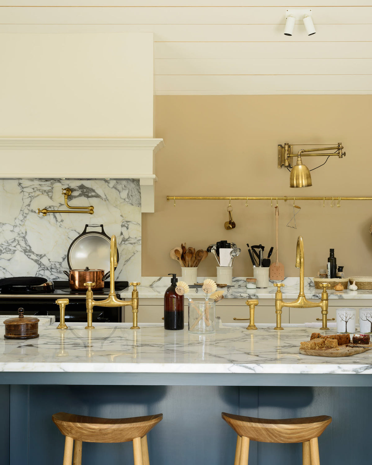 A grand mushroom-coloured classic English kitchen features a cooker run with a suspended chimney breast, a large kitchen island, two sets of sinks, aged brass Ionian taps, and an Arabescato marble worktop. Beautifully decorated with a aged brass hanging rail, bum stools, simple ceramics, a brass folded wall lamp, and a large collection of kitchen utensils, this space creates a humble family atmosphere despite its luxurious design.