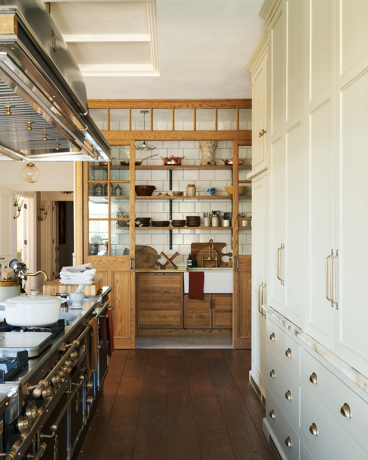 Looking down the grand kitchen with a high panelled ceiling, traditional glossy white tiled walls, and a huge bank of creamy mushroom full-height classic cupboards is a large haberdasher’s pantry. It’s complete with its own haberdasher’s sink run topped with a marble worktop, large shelves, and an assorted collection of ceramic plates, bowls, pots, pans, and silverware.