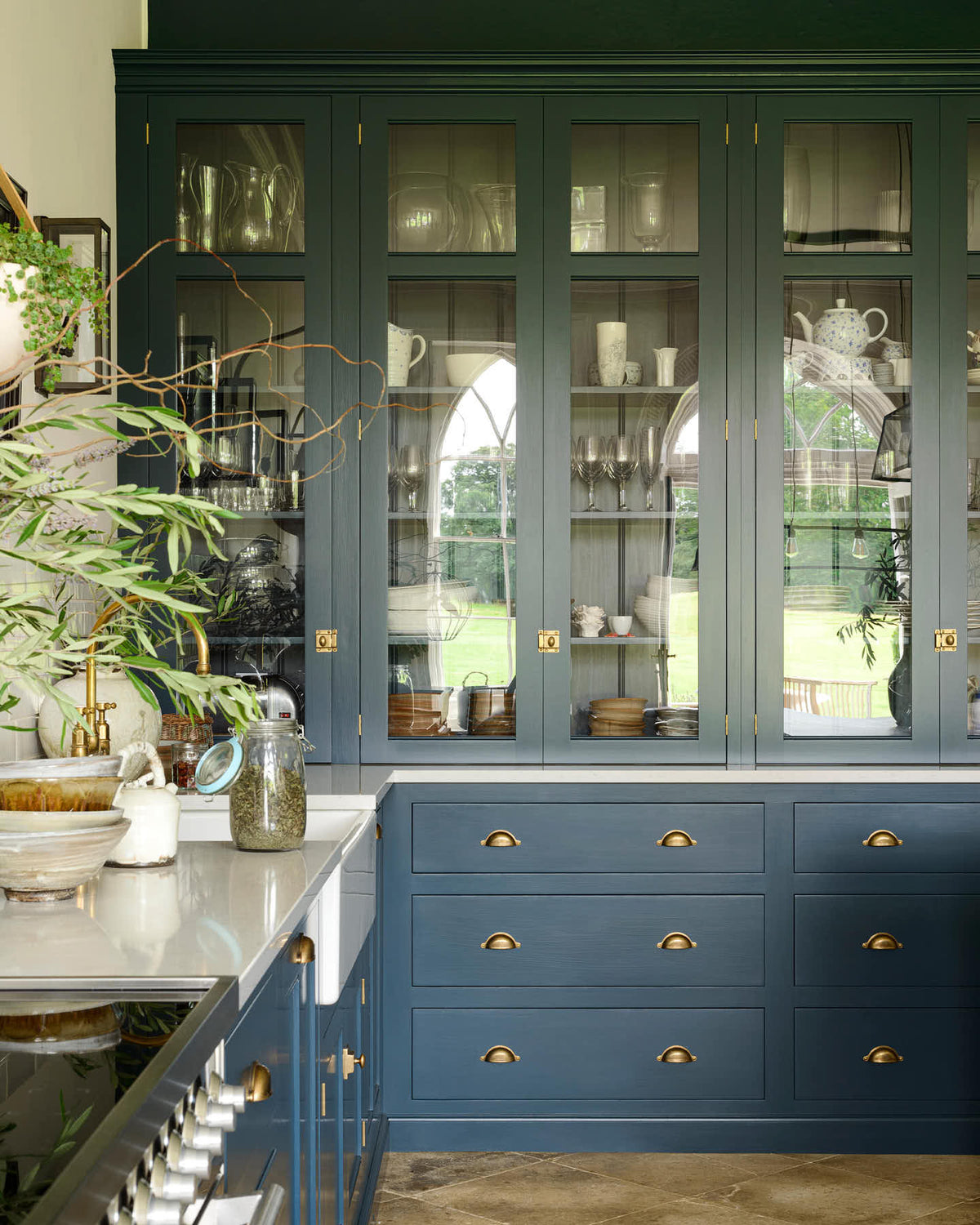 A classic English, Bond Street blue kitchen features a cooker and sink run with a Silestone worktop, glazed counter cabinets, a double sink, and an aged brass Ionian tap. The renovated medieval house is elegantly adorned with modern still-life paintings, artisan ceramics, and vibrant green plants, seamlessly blending the historical with the contemporary.