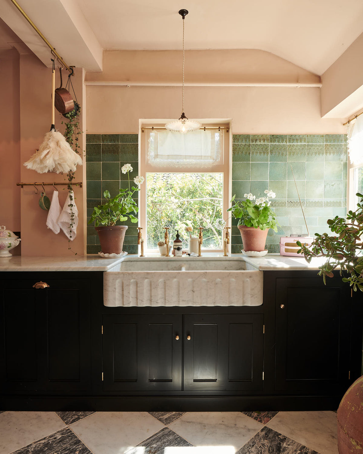 A closer look at the bright peach-coloured kitchen reveals a Carrara marble worktop with a double fluted sink and twin aged brass Mayan taps. Vintage teal lace-market splashback tiles add character, paired with a small brass rail holding a teal teacup and tea towels. The space is styled with an eclectic mix of vintage pieces, including a wall-mounted candle holder, a radio, a gold-framed painting of a tomato, and potted plants dotted across the counter.
