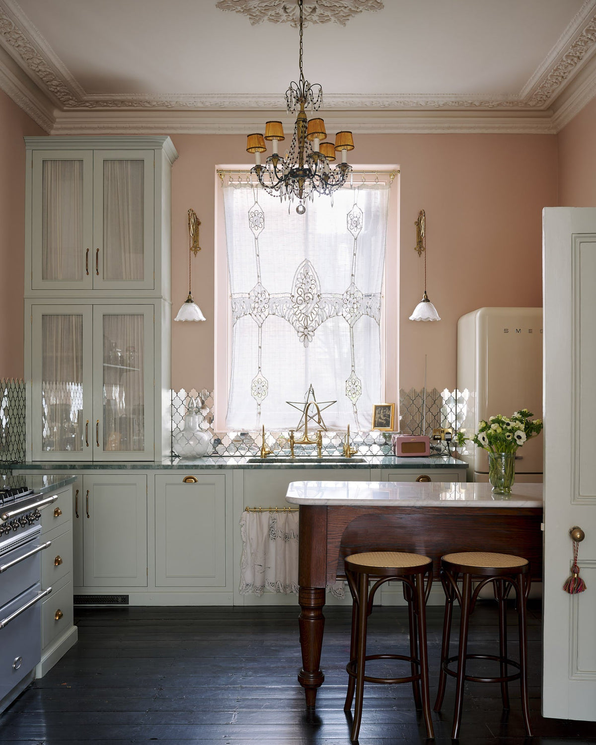 A light pastel-blue classic kitchen and social area features a cooker and sink run with marble worktops, an heirloom dairy table, and Bentwood café stools—colours that oddly fit perfectly into the plaster-pink room, creating a glamorous yet eccentric vibe. The London apartment has high ceilings, worn black painted floorboards, small decorative splash back mirrors, intricate cornices, and is styled with the customer's collection of antiques.