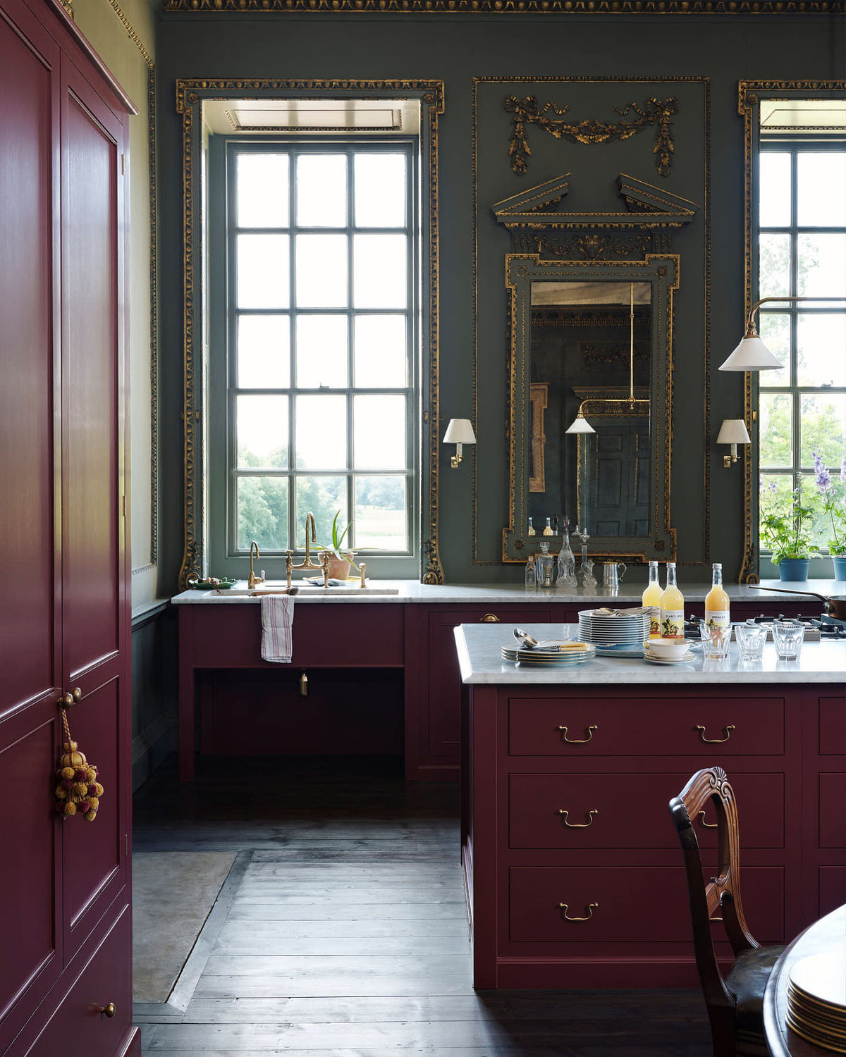 A corner of a grand, refectory red classic English kitchen featuring a sink run and cooker with Carrara marble worktop, a kitchen island, and a tall pantry cupboard. Place upon a tall, magnificent open space with deep olive green walls with ornate gold gilding, and beautifully decorated with a Gaselier light, artisan ceramics, and vintage furniture, creating a splendidly royal and practical use of space.