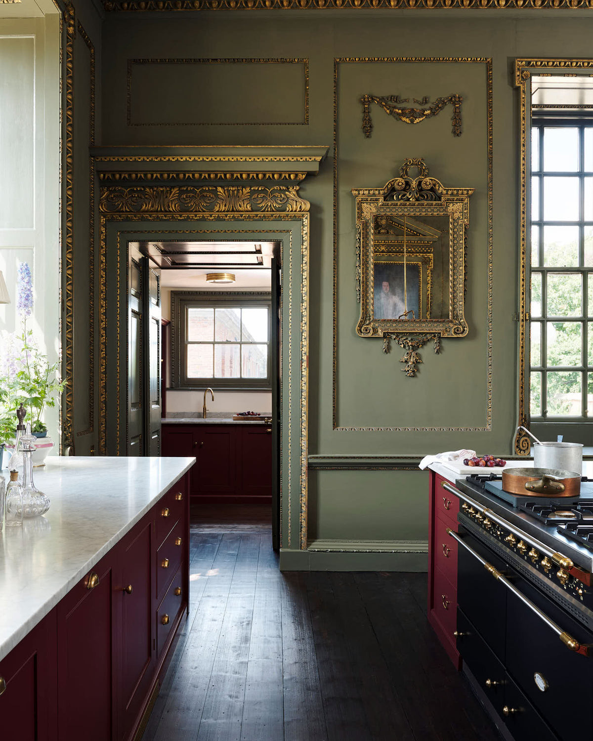 A grand, refectory red classic English kitchen featuring a sink run with Carrara marble worktop, a kitchen island, and a small scullery. Place upon a tall, magnificent open space with deep olive green walls with ornate gold gilding, and beautifully decorated with a Gaselier light, artisan ceramics, and vintage furniture, creating a splendidly royal and practical use of space.