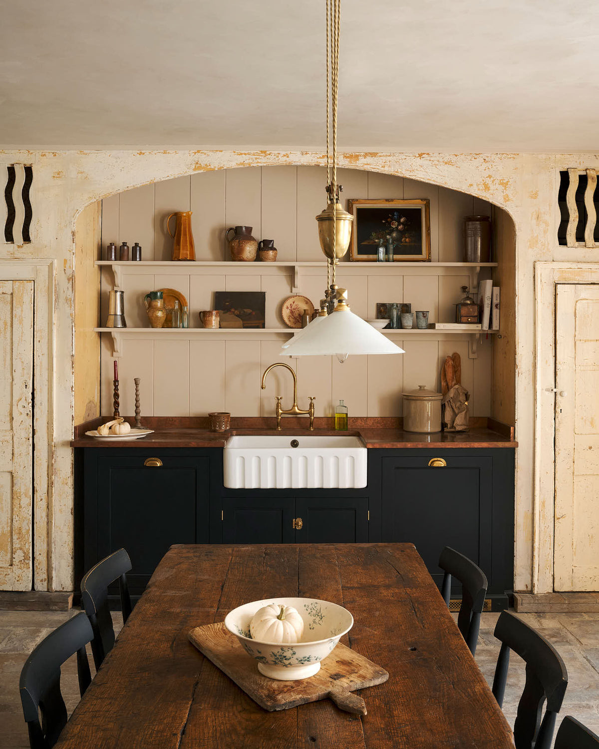 A rustic basement kitchen with sandy-coloured walls and ceiling, original Georgian detailing, cream doors and panelling with chipped paintwork and a worn stone floor. An old banquet-style table sits in the middle of the room pointing at a fluted Belfast sink and an aged brass tap below shelves decorated with vintage bric-a-brac.