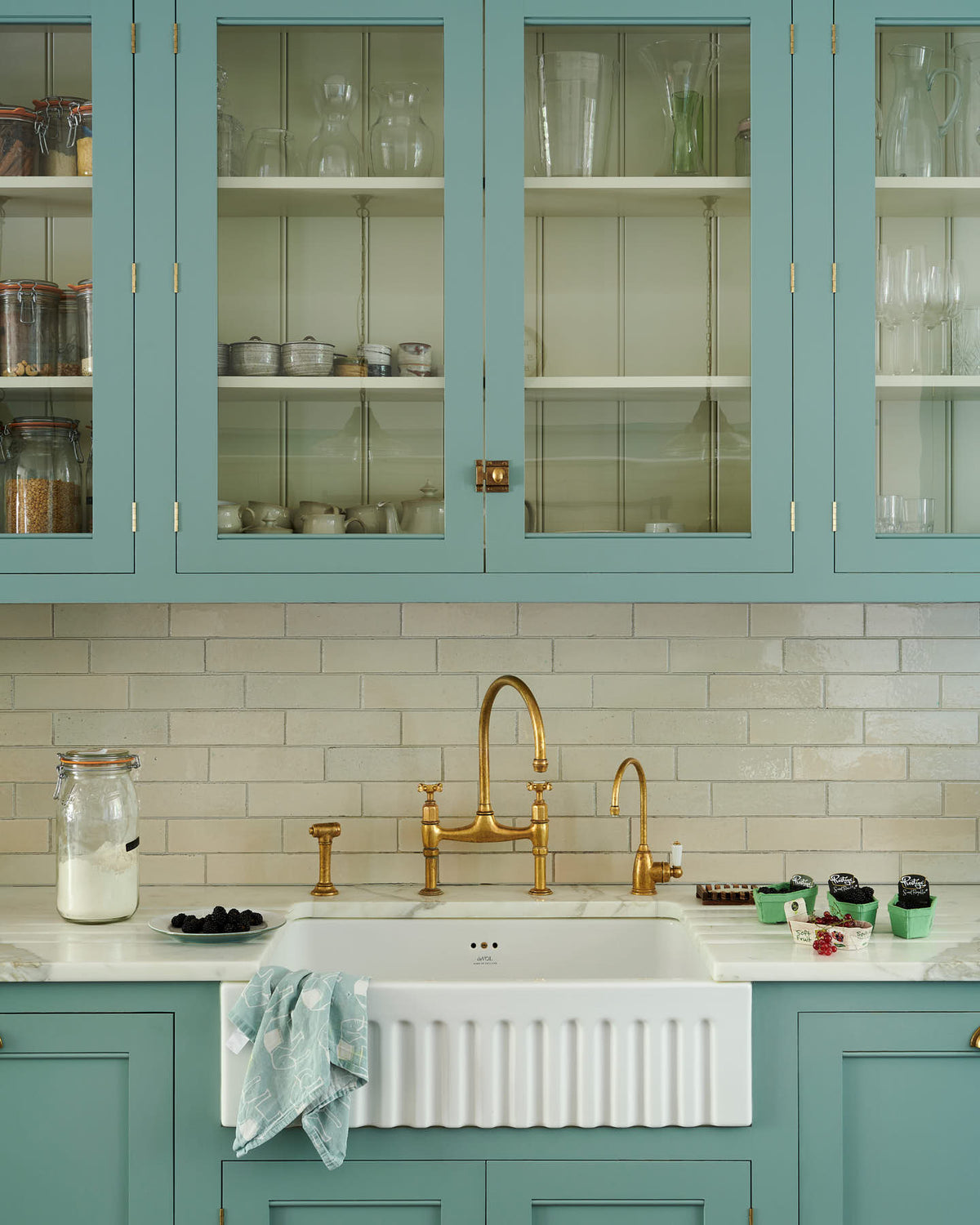 A spacious yet modest Trinity Blue classic English kitchen features a Silestone worktop sink run with a single fluted Belfast sink and aged brass Ionian tap, glazed cupboards, and a kitchen island with a copper sink—all topped off with a white porcelain splashback. Lovingly decorated with a silver juicer, papayas, live basil, berries, dried ingredients in glass jars, the room feels humble, warm, and inviting.