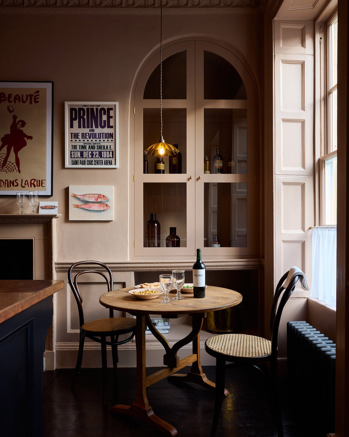A quiet corner of a bold and dramatic Haberdasher’s kitchen features a deVOL petite vineyard table, Bentwood café chairs, and a petite frilly chartreuse light. Adorned with a delicious bottle of wine, drinking glasses, hors d’oeuvres, quirky framed illustrations and posters, and labelled glass jars, this space creates a local bistro bar atmosphere in Manhattan.
