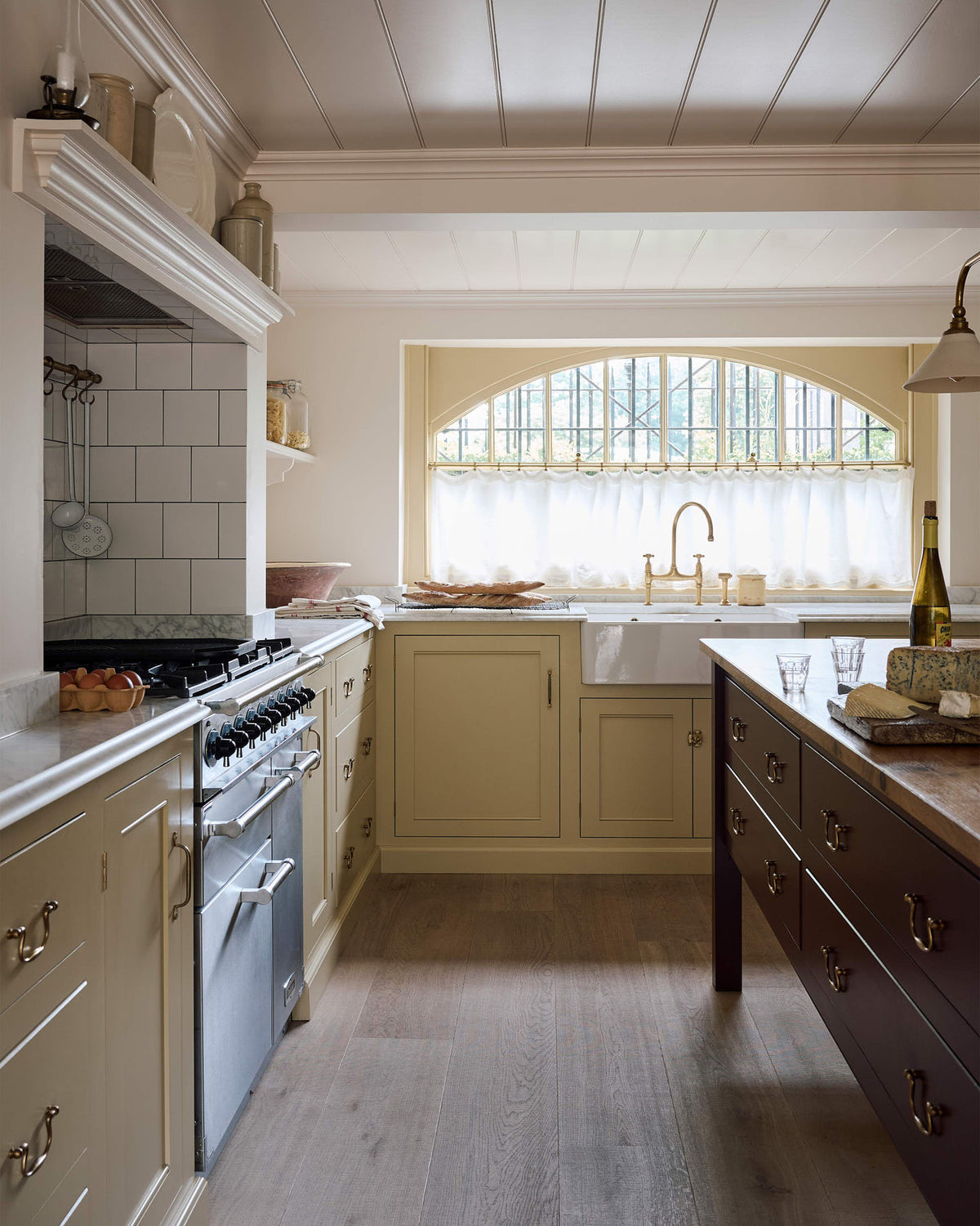 A warm, inviting, muted yellow classic English kitchen features an L-shaped cooker and sink run with a Carrara marble worktop and a large freestanding maroon prep table with a solid oak worktop. The double Belfast sink with an aged brass Ionian tap sits in front of a semi-circular window with an aged brass curtain rail at the bottom half, allowing privacy and ample natural light to brighten the room.