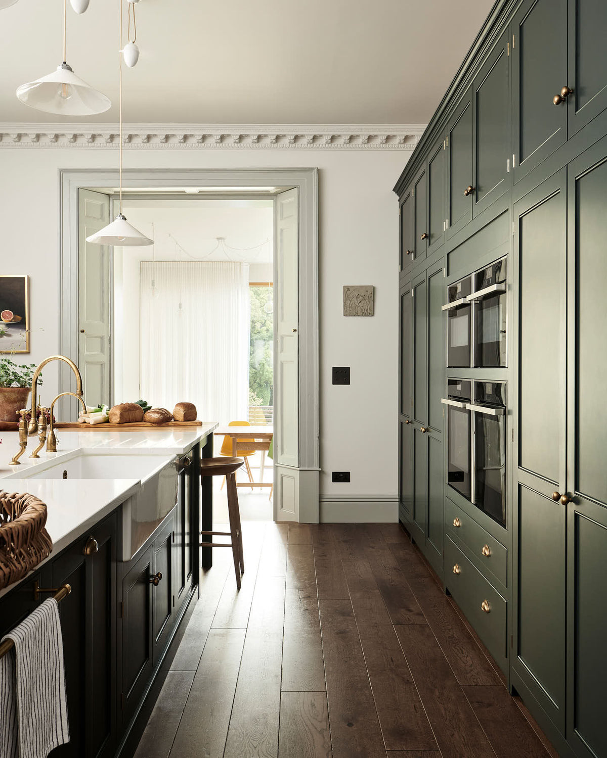 Looking between a large full-height pantry cupboard, oven cabinets, fridge/freezer cupboard, and a long kitchen island with a double sink, an aged brass Ionian tap in the grand bakehouse green classic English kitchen. Adorned with loaves of bread, leeks, potted plants, and still life paintings; the room feels homey and welcoming despite its large and decorative appearance.