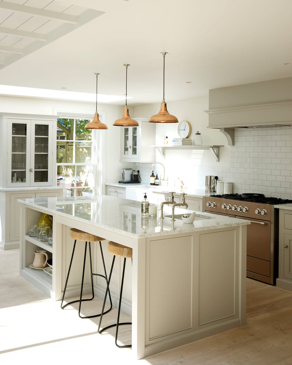 This simple and stylish classic English kitchen features a sink and cooker run, a suspended false chimney breast, a large pantry cupboard, and a kitchen island topped with Carrara marble. Beautifully decorated with light copper pendant lights, a silver toaster, a silver kettle, white ceramics, and white splashback tiles, it creates a functional and beautiful kitchen space.