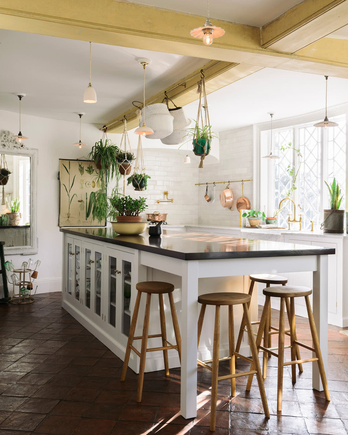 The white linen, classic English kitchen features an L-shaped sink and cooker run with Carrara marble worktop, a large single Carrara marble sink, an aged brass Ionian tap, and white crackle metro splash back tiles. In the centre is a large kitchen island with a solid oak worktop, breakfast nook and Helen stools. Lush green hanging plants and white porcelain lights, with the original yellow beam, enhance the bright and cheerful atmosphere, making the kitchen lively and inviting.