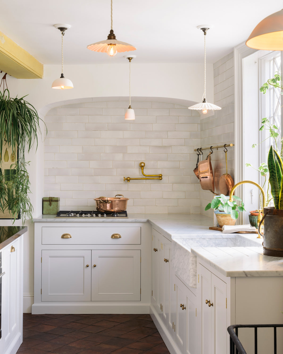 The white linen, classic English kitchen features an L-shaped sink and cooker run with Carrara marble worktop, a large single Carrara marble sink, an aged brass Ionian tap, and white crackle metro splash back tiles. In the centre is a large kitchen island with a solid oak worktop, breakfast nook and Helen stools. Lush green hanging plants and white porcelain lights, with the original yellow beam, enhance the bright and cheerful atmosphere, making the kitchen lively and inviting.