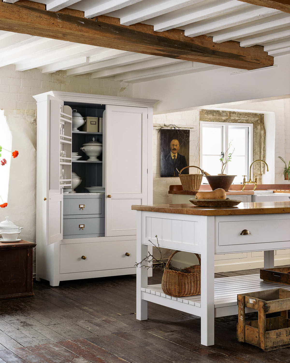 An angled view of a beautiful white linen kitchen featuring a kitchen run with an aged brass Ionian tap, freestanding table, a pantry cupboard, and a collection of vintage furniture and items reflecting on the well-reserved historical space with its original plastered walls, wooden beams and oak floors.