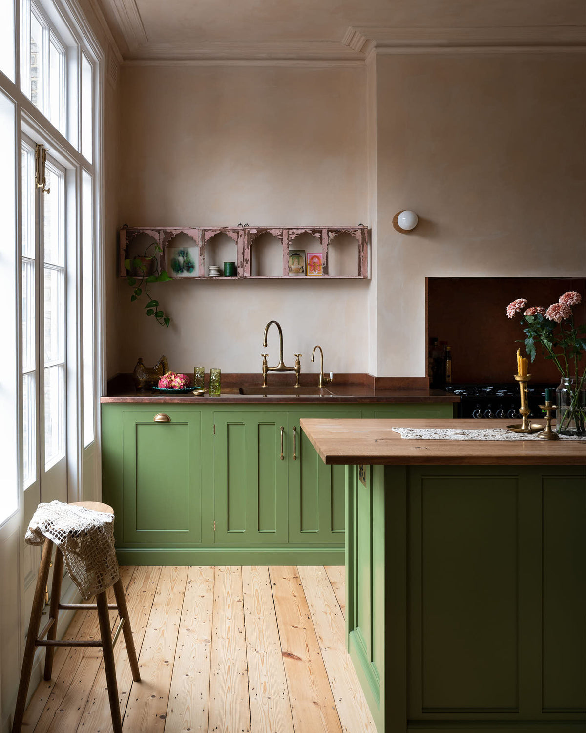 A romantic, decorative, Parisian-inspired bean green classic English kitchen featuring an L-shaped sink and cooker run with an aged copper worktop, an aged brass Ionian tap, and an oak worktop kitchen island. Above the sink, a pink vintage-framed shelf is adorned with small colourful abstract prints, lace runners, a bouquet of blush-brown chrysanthemums, and a strikingly red plate of dragon fruits.