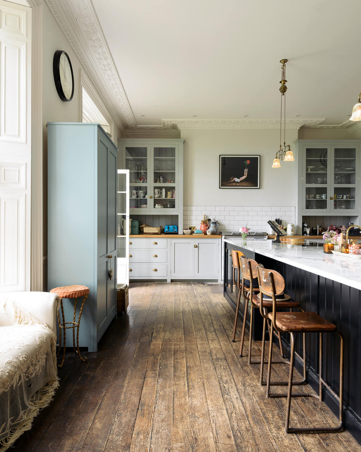 A tall-ceilinged classic English kitchen centres around a wide painter’s black island with an aged brass Ionian tap, marble worktop, and glazed base cabinets. A cosy breakfast nook with vintage stools sits opposite an off-white cooker run and matching wall cabinets. The space feels artfully lived-in—funky paintings, and kitchen essentials tell the story of a creative, full-of-life family.