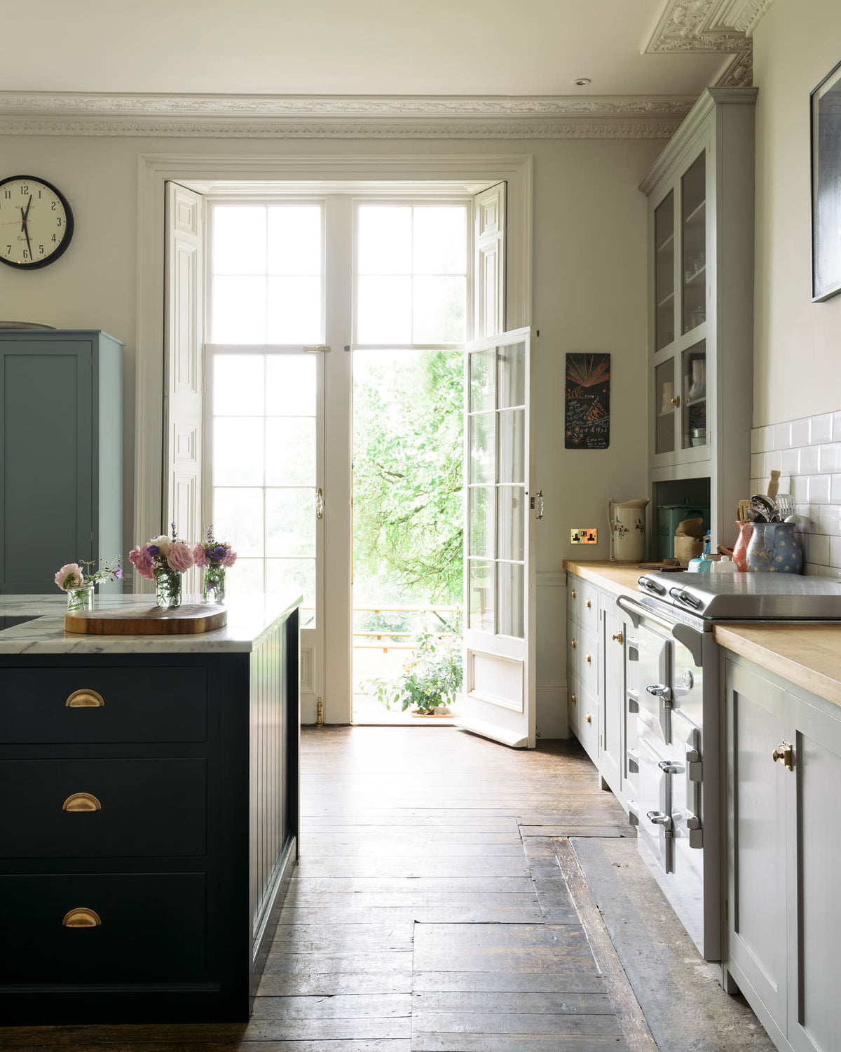 One side of a French door is left open to the lush green garden, allowing bright white natural light to pour into the tall-ceilinged, classic English kitchen centred around a wide painter’s black island with a marble worktop, and an off-white cooker run with matching wall cabinets and white porcelain splashback tiles.