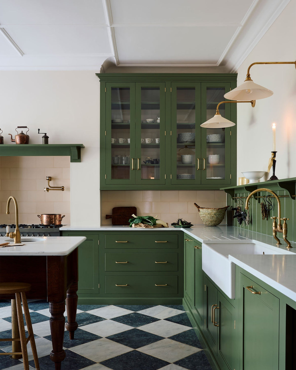 A spacious and elegant Wilkes green classic English kitchen features a long L-shaped cooker and sink run with a silestone worktop, glazed wall cupboards, shelves, and an Heirloom dairy table. Adorned with classic marble chequered-board flooring, Heirloom task lights,  cream subway splash back tiles, copper pots, and luscious green vegetables, it creates a classic and stylish kitchen.
