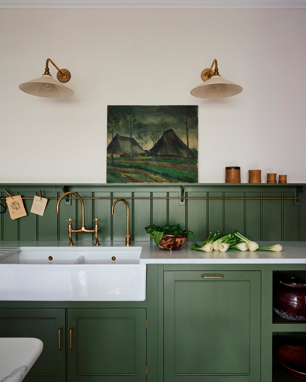 A closer look at a spacious and elegant Wilkes green classic English kitchen featuring a sink run with a double Belfast sink, an aged brass Ionian tap, a low tongue-and-groove panel, and a long shelf with hanging rail. Adorned with vintage containers, Heirloom task lights, a moody landscape painting, and luscious green vegetables, it creates a classic and stylish kitchen.