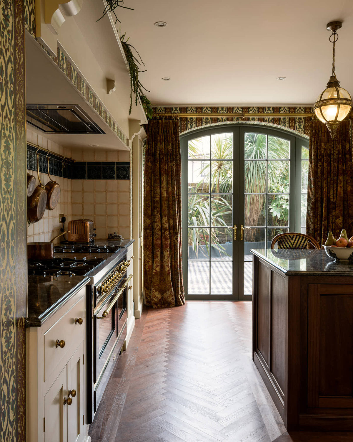 This grand and luxurious classic English kitchen features a cooker run set into the chimney breast, a large kitchen island topped with Volga blue granite, a single copper sink, an aged brass Ionian tap, and free-standing cupboards. Lavishly decorated with intricately patterned wallpaper, reclaimed splashback tiles, and hanging lantern pendant lights, it creates a royal yet comfortable and quirky space.